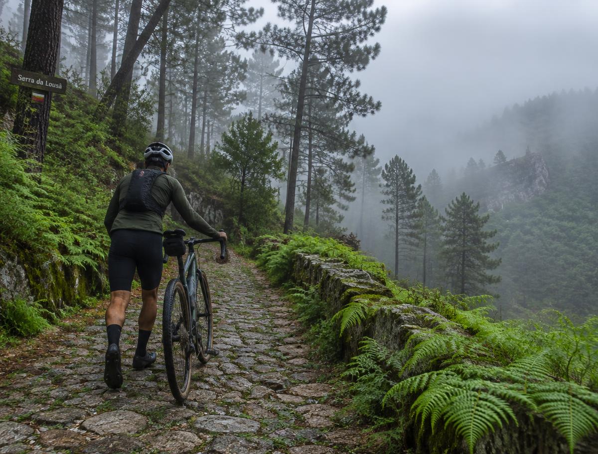 Cyclist walking a stone lane through the misty mountain forests of the Serra da Lousã